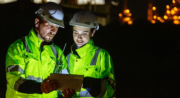 Two contractors in hardhats look at a clipboard at night