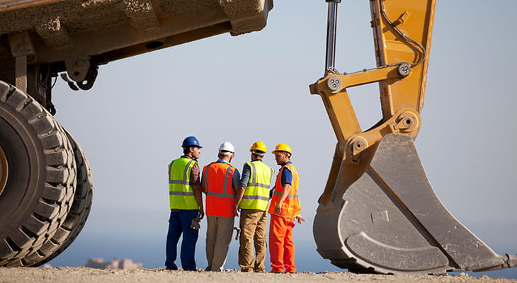 Four construction workers stand under a big excavator in a remote site